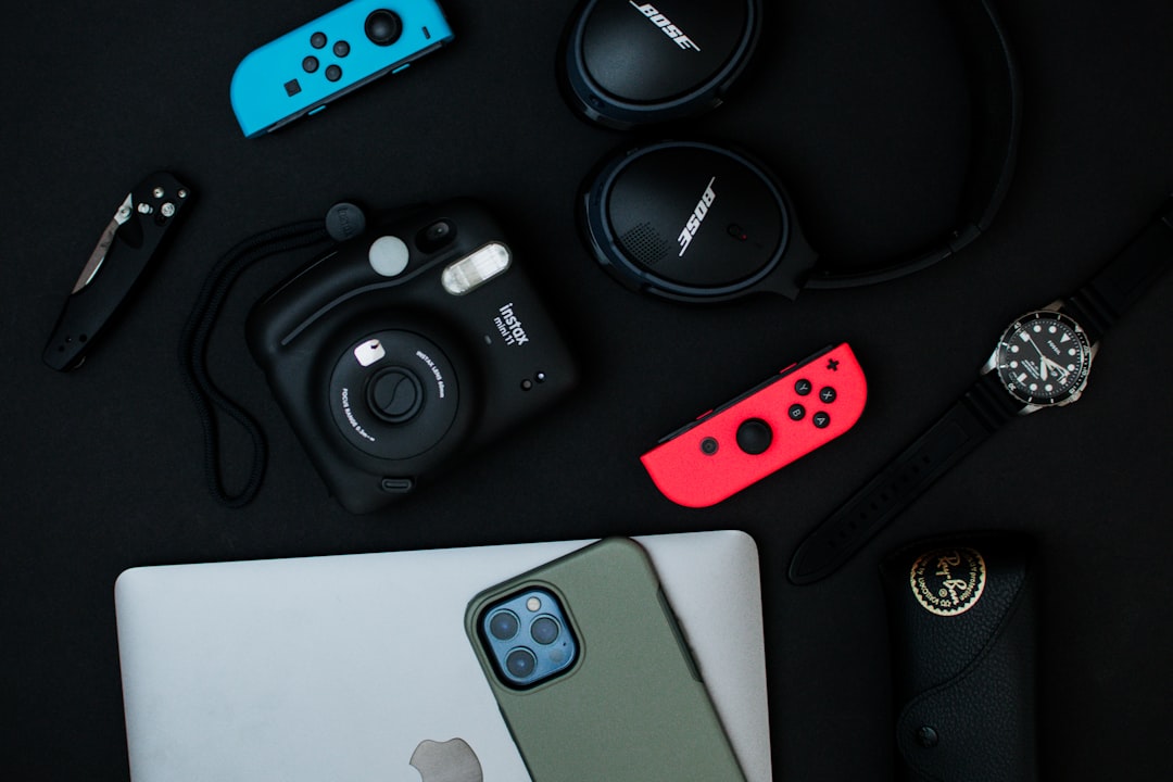 Flat lay headphones, watch, sunglasses, knife, laptop, phone, joycons, and camera on black backdrop
